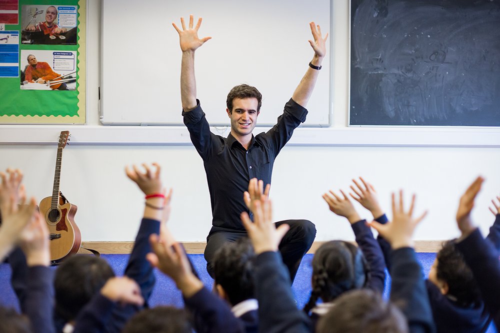 A smiling instructor leads a lively music class, arms raised high, mirroring the enthusiastic response of young students who participate with equal excitement. A guitar rests beside the blackboard, hinting at the musical journey they're on together.