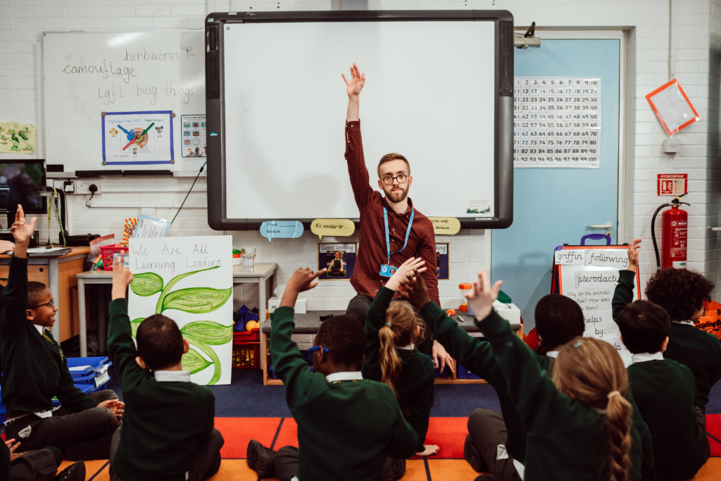 A classroom scene captures a group of enthusiastic students seated on the floor, raising their hands to answer a question posed by their teacher, who stands at the front with his hand also raised, signaling for a response. The environment is vibrant with educational materials, including a whiteboard, posters, and books, fostering an interactive and engaging learning experience.