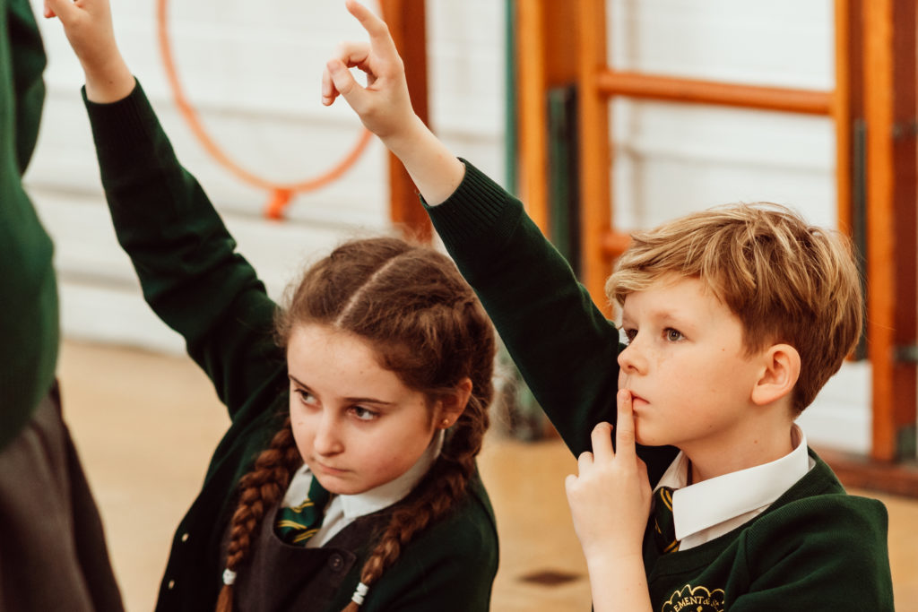 Two schoolchildren in uniform are engaged in a classroom activity, with one child thoughtfully raising a finger to their face, and the other with an arm raised, possibly in response to a question or as part of an interactive lesson. Their expressions suggest concentration and curiosity, capturing a moment of learning and participation.