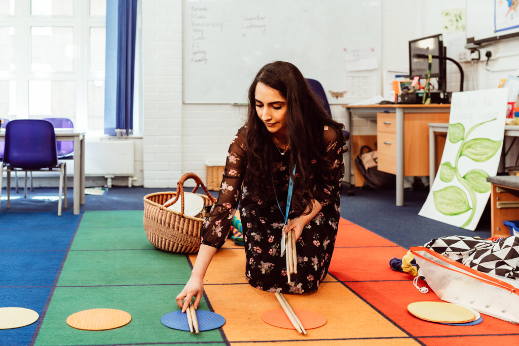 A teacher kneels on a colorful classroom carpet, arranging musical instruments next to vibrant circular mats, preparing for an engaging and interactive music lesson that promises to captivate and educate young learners. The background reveals a bright and inviting educational environment with a whiteboard listing musical terms, fostering a creative and nurturing atmosphere for learning.
