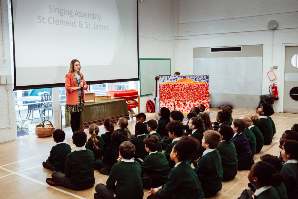 A group of schoolchildren in green uniforms sits attentively on the floor of a spacious room, their focus directed towards a woman standing in front of a large screen displaying the words "Singing Assembly St Clement & St James." The woman, dressed in a red blazer and patterned dress, appears to be leading the assembly, with a sense of calm engagement shared between her and the students. The setting suggests an interactive and educational environment, likely filled with music and learning.