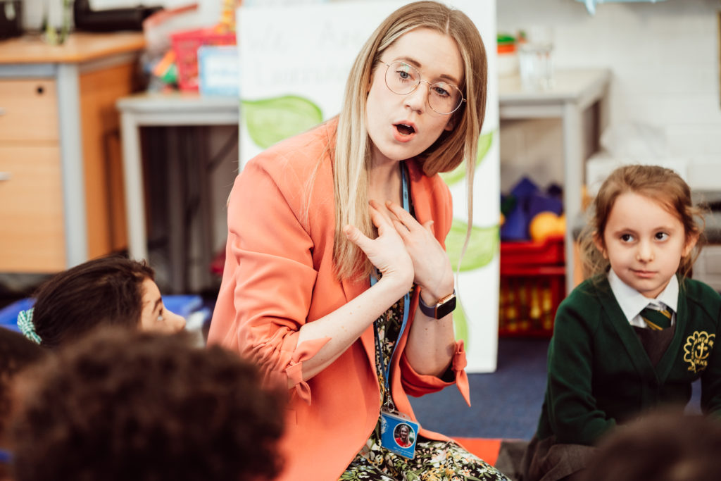 A teacher, animatedly engaged in a classroom activity, captures the attention of her young students with a look of surprise and curiosity on her face, illustrating the dynamic and interactive nature of learning. The children, dressed in school uniforms, watch intently, suggesting a moment of discovery or the introduction of a new concept. The classroom setting, with educational materials in the background, underscores the learning environment.