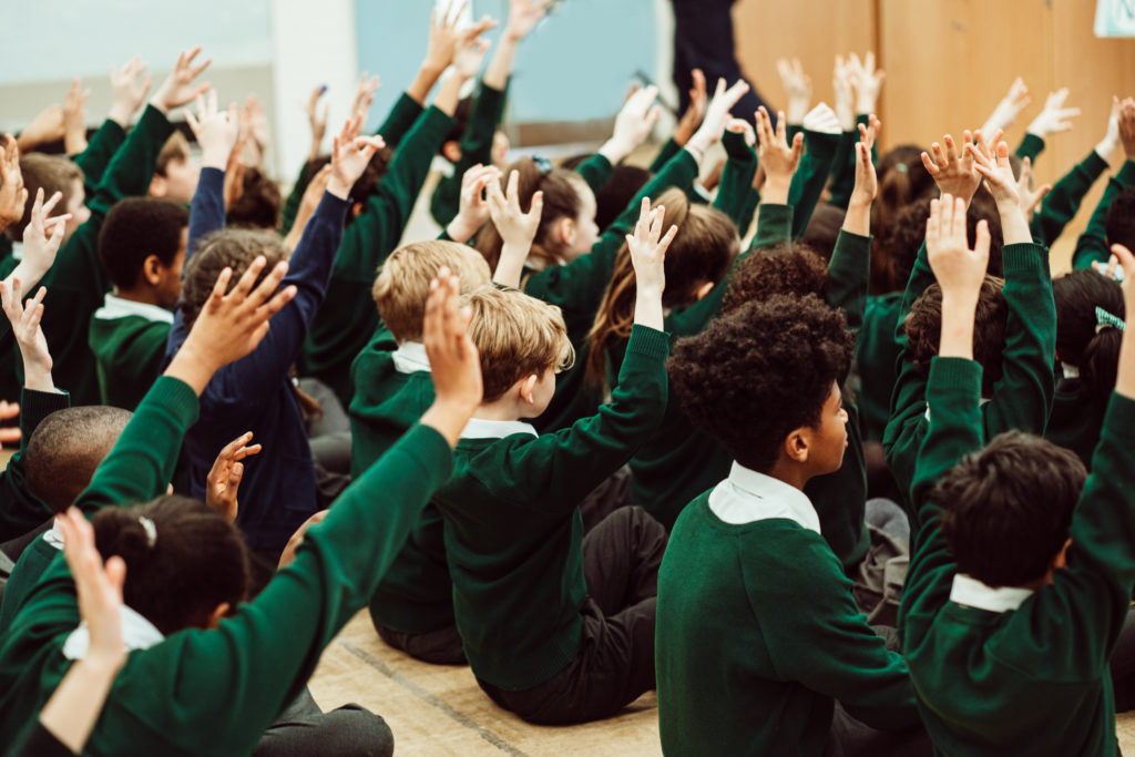 A group of enthusiastic schoolchildren, dressed in green uniforms, are seated on the floor with their hands raised high in a gesture of participation. The atmosphere is one of active engagement, likely in response to a teacher's question or as part of an interactive learning activity, capturing a moment of collective eagerness and curiosity among the students.