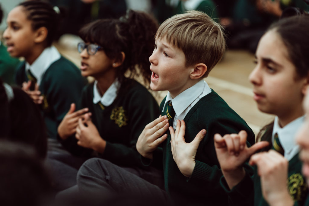 A group of schoolchildren in green uniforms are engaged in a singing activity, with one boy in the foreground singing passionately and gesturing with his hands, surrounded by his classmates who are also participating with enthusiasm. The atmosphere is one of focus and enjoyment as they partake in this musical learning experience.