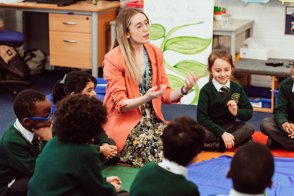 A classroom comes alive with a group of attentive primary school students sitting on the floor, encircling a teacher who is engaging them with a hands-on activity. The teacher, dressed in a bright coral blazer, gestures expressively, fostering a dynamic learning environment. The children, in green uniforms, appear focused and curious, contributing to the interactive educational experience. A poster in the background reads "We Are All Learning Leaders," reinforcing the collaborative atmosphere.