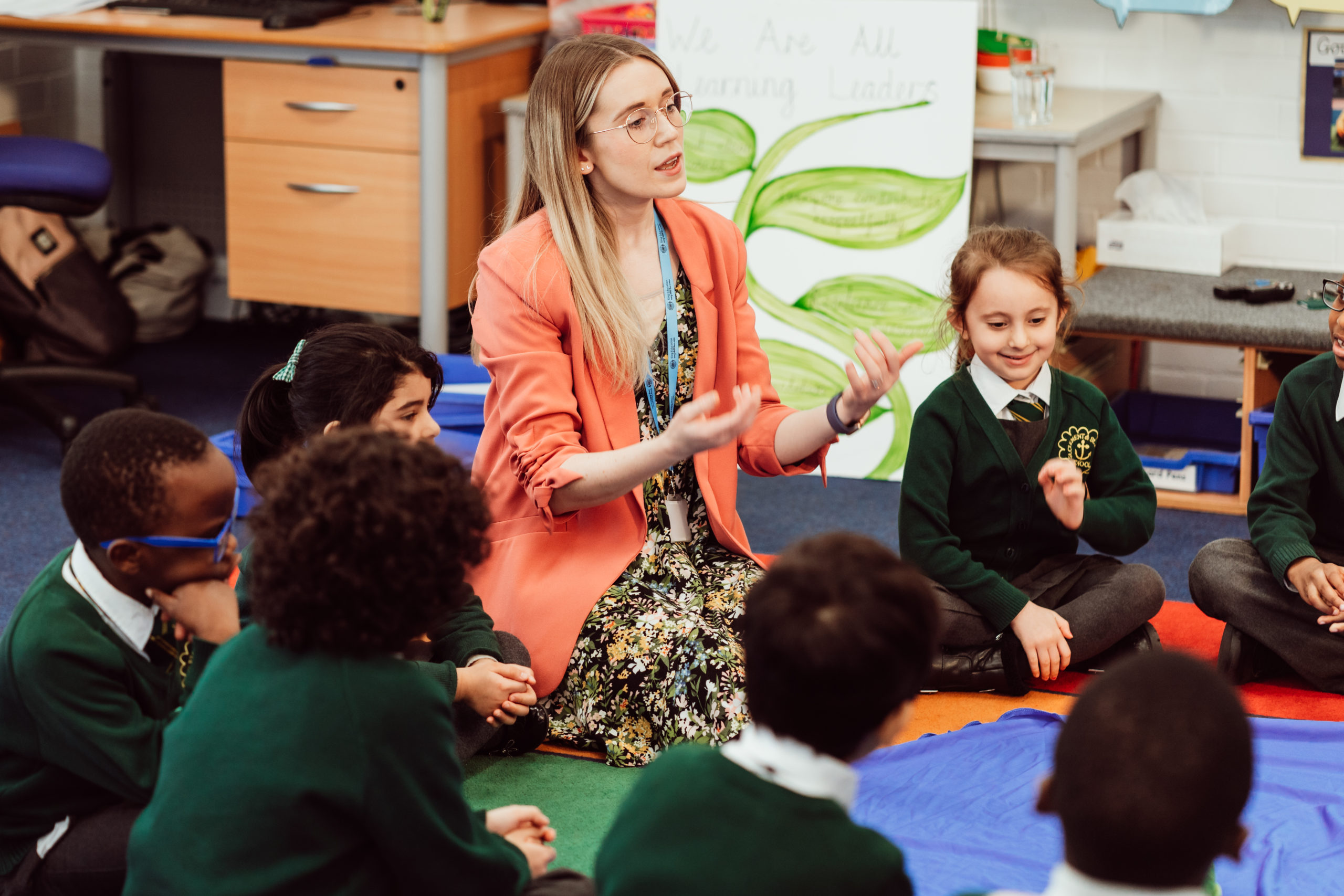 A classroom comes alive with a group of attentive primary school students sitting on the floor, encircling a teacher who is engaging them with a hands-on activity. The teacher, dressed in a bright coral blazer, gestures expressively, fostering a dynamic learning environment. The children, in green uniforms, appear focused and curious, contributing to the interactive educational experience. A poster in the background reads "We Are All Learning Leaders," reinforcing the collaborative atmosphere.