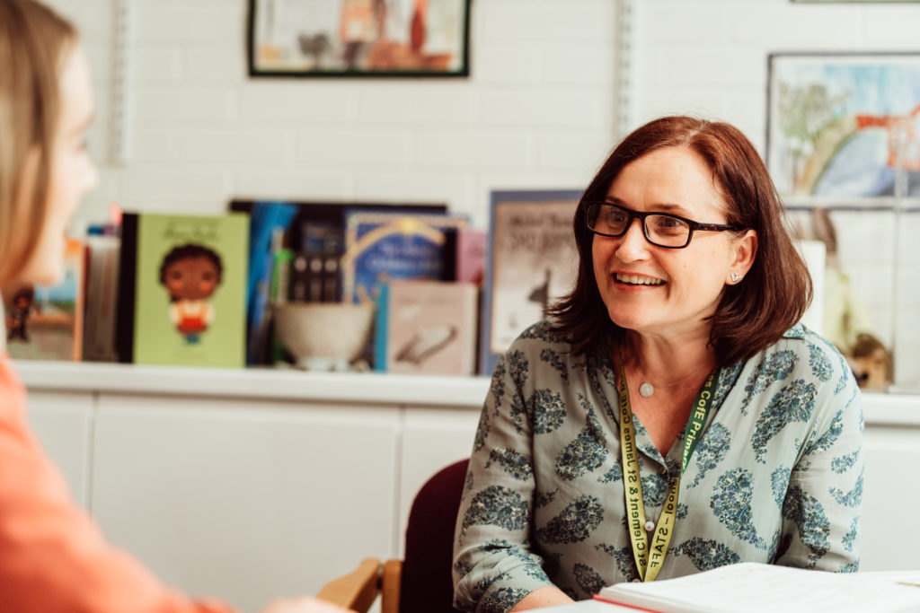 A smiling woman with glasses, wearing a floral blouse and a lanyard, is engaged in a conversation with a person whose back is to the camera. They are seated indoors, with a bookshelf filled with various books and educational materials in the background, creating a warm and inviting educational environment.