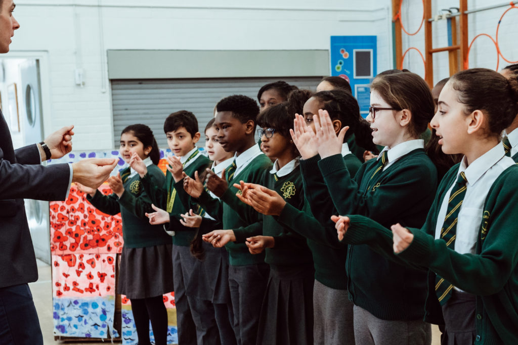 A group of enthusiastic schoolchildren in uniform are actively participating in a music class, attentively following the hand gestures of their instructor, who is leading them with visible engagement. The children appear focused and joyful as they engage in the learning activity, demonstrating the interactive and vibrant nature of music education.