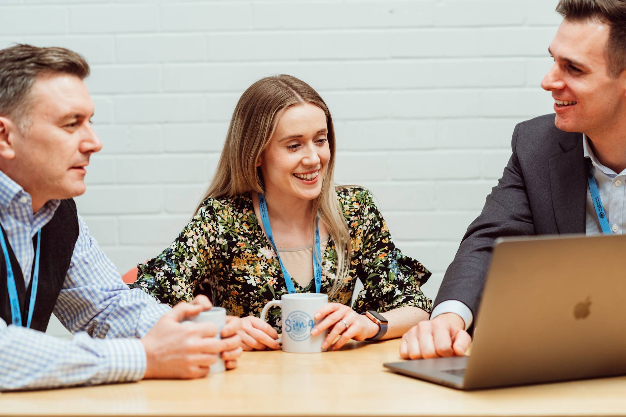 Three professionals are engaged in a lively discussion around a wooden table, with one woman and two men smiling and sharing ideas. The woman, in the center, holds a mug with the Sing Education logo, while a laptop is open in front of the man on the right, suggesting a collaborative work environment. The casual yet focused atmosphere indicates a productive meeting, with the white brick wall in the background providing a modern, relaxed setting.