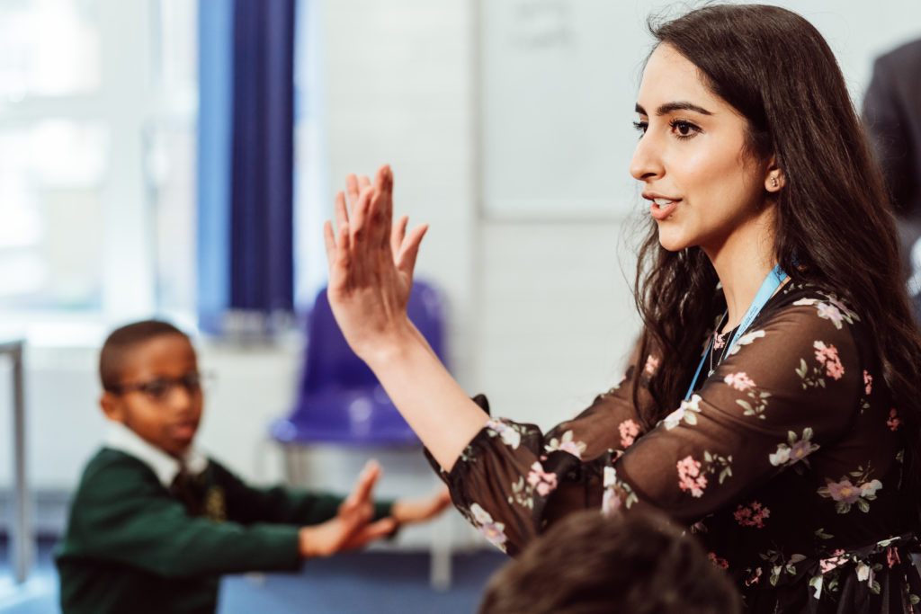 A focused teacher with long hair and a floral dress is engaging with her students in a classroom setting, using hand gestures to illustrate a point or direct the activity, while a young student in a green sweater attentively participates in the foreground. The atmosphere suggests an interactive and dynamic learning experience.