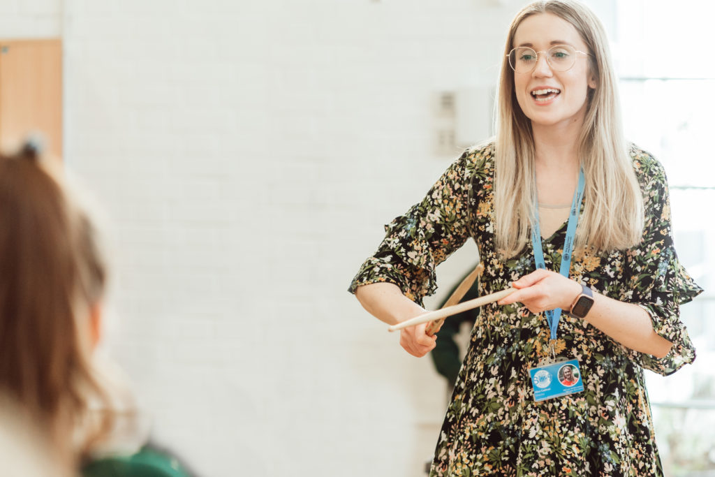 A smiling woman in a floral dress and glasses, holding a conductor's baton, leads a music class, engaging with a student who is partially visible in the foreground. Her lanyard and badge suggest she is a teacher or instructor, and the bright, airy room enhances the lively educational atmosphere.