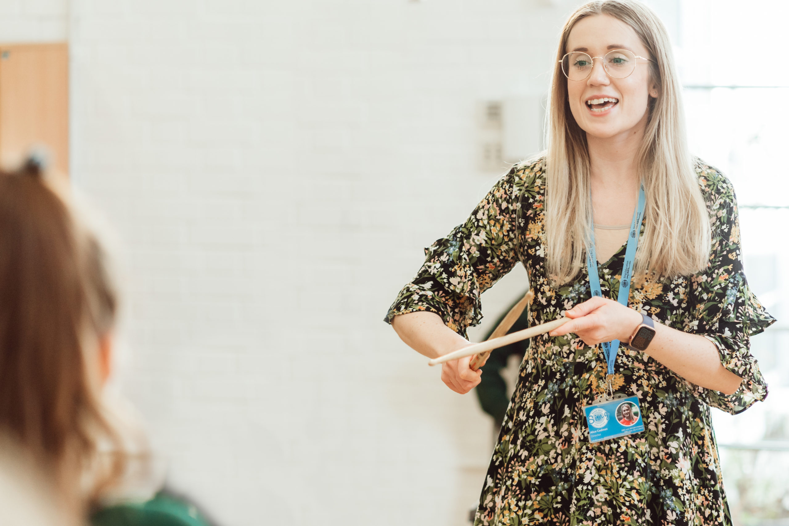 A smiling woman in a floral dress and glasses, holding a conductor's baton, leads a music class, engaging with a student who is partially visible in the foreground. Her lanyard and badge suggest she is a teacher or instructor, and the bright, airy room enhances the lively educational atmosphere.