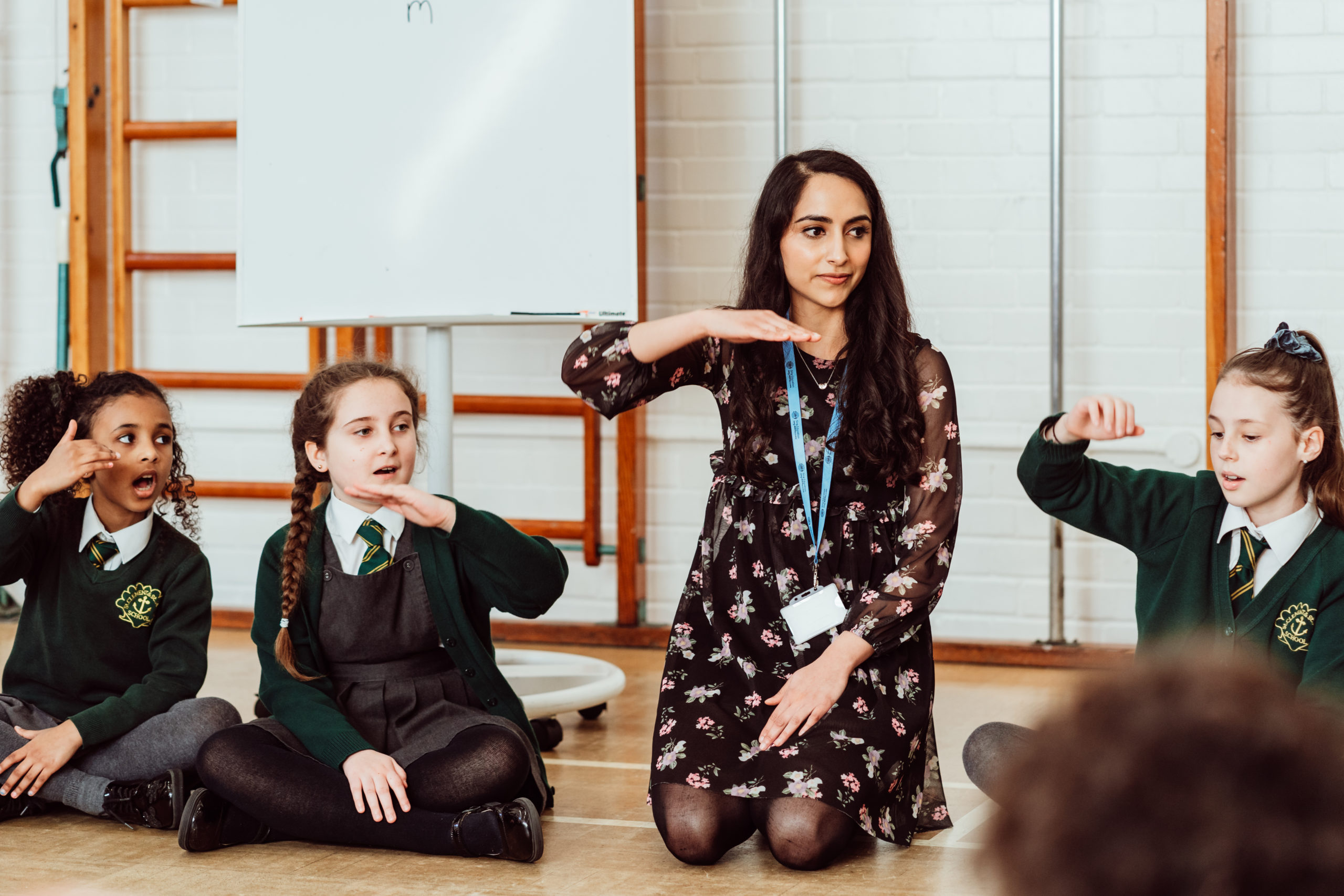 A teacher and three students are engaged in a lively classroom activity; the teacher is kneeling and demonstrating a hand gesture, while the students, seated on the floor, mimic the motion with expressions of focus and enjoyment. The classroom setting includes a whiteboard in the background, suggesting an interactive learning environment.