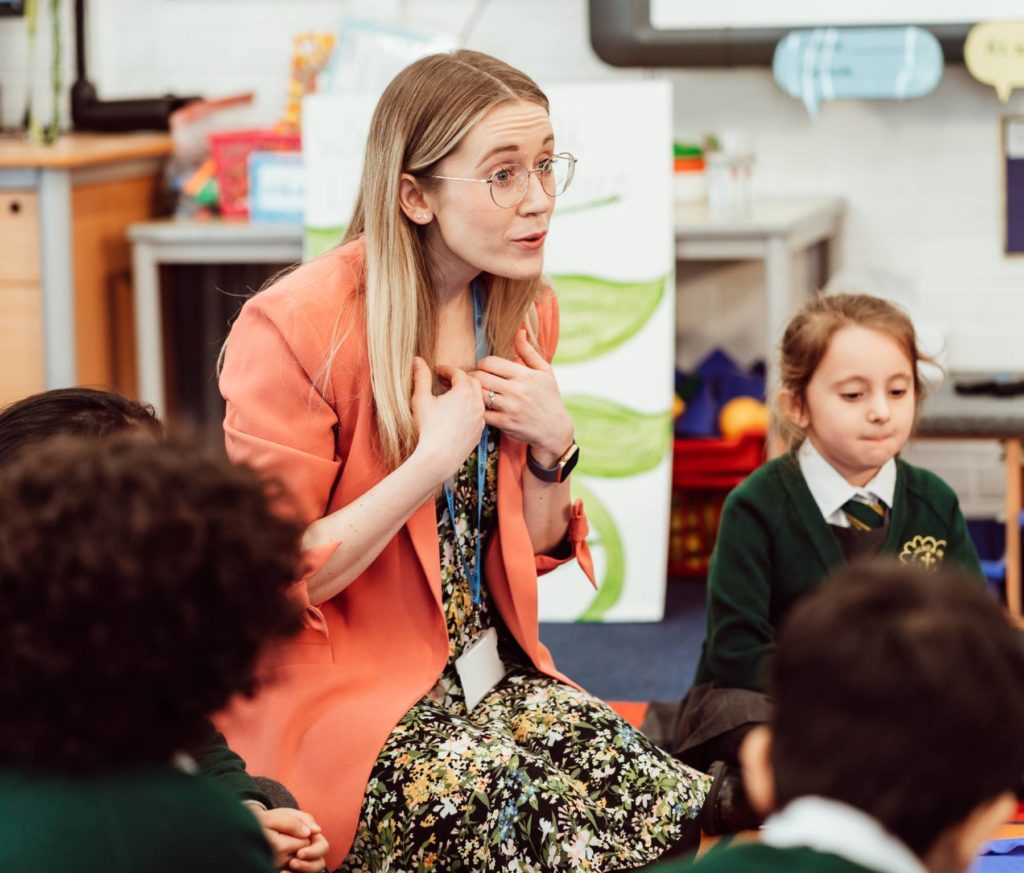 A teacher, dressed in a floral dress and coral blazer, sits cross-legged and engages with young students in a colorful classroom setting. She gestures to herself with an animated expression, captivating the children who are attentively listening to her, fostering a lively and interactive learning environment.