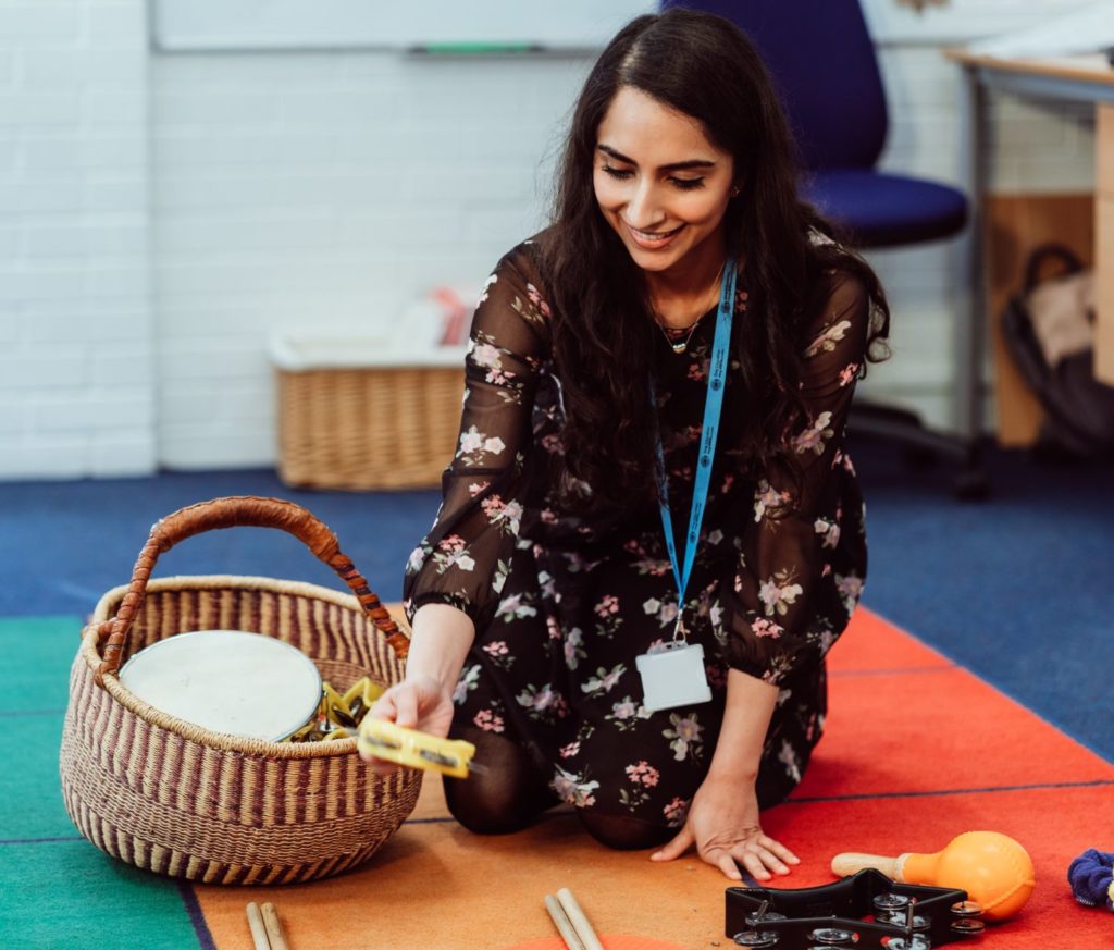 A smiling educator kneels on a colorful classroom rug, reaching into a woven basket filled with musical instruments like tambourines and shakers, creating an inviting and interactive learning environment.