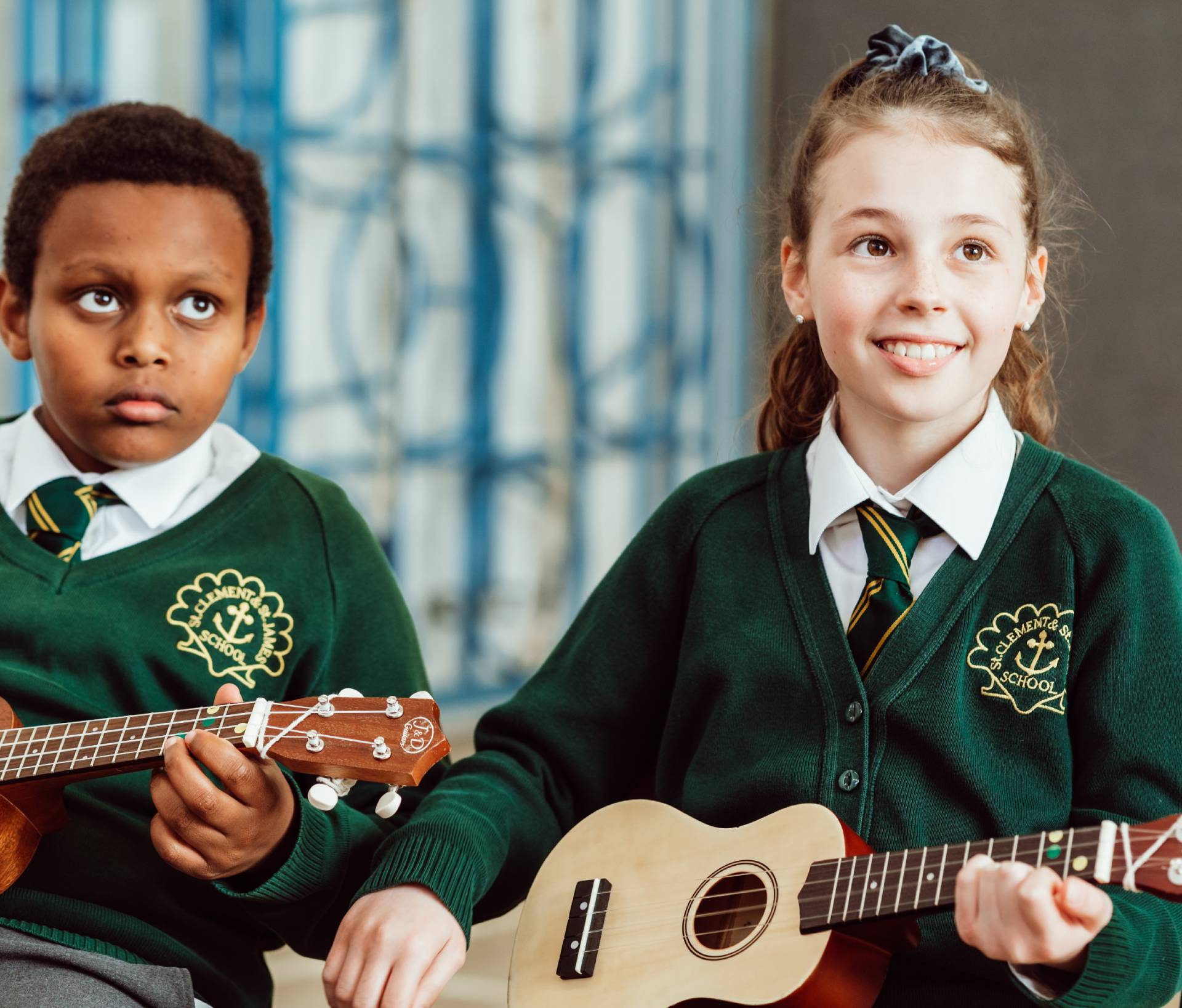 Two schoolchildren in green sweaters with embroidered school crests are seated with ukuleles, the girl smiling brightly as she looks ahead, while the boy appears focused on his instrument, both poised to play against a backdrop of a patterned blue curtain. Their engagement suggests a lively music lesson in progress, highlighting the joy and concentration that comes with learning an instrument.