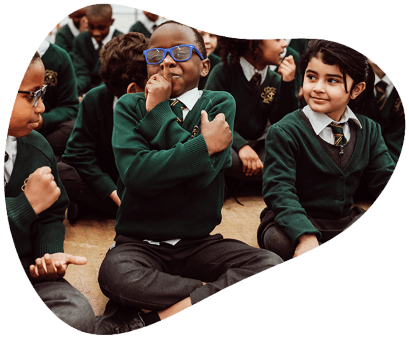 A group of schoolchildren in green sweaters and ties are seated on the floor, with one boy in the foreground wearing blue glasses, enthusiastically participating in a classroom activity. The children appear engaged and focused, with some holding their hands to their chests, suggesting a collective learning experience or musical activity that promotes involvement and enjoyment.