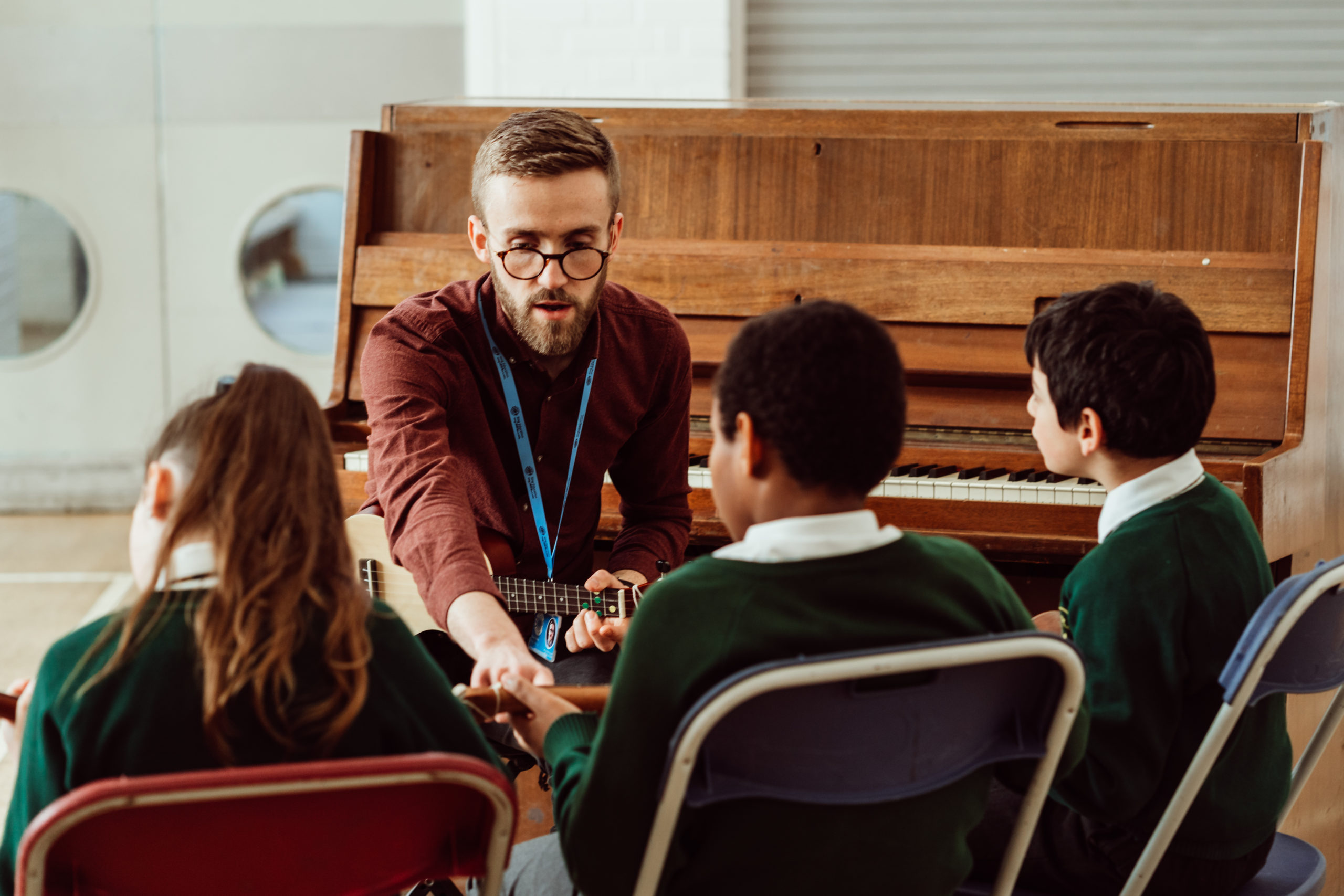 A music teacher with a beard and glasses is seated, playing a guitar, surrounded by three attentive children in school uniforms. They are engaged in a music lesson, with a piano in the background, suggesting a creative learning environment where the teacher is actively involving the students in the musical experience. The atmosphere is one of focus and enjoyment as the group explores the world of music together.