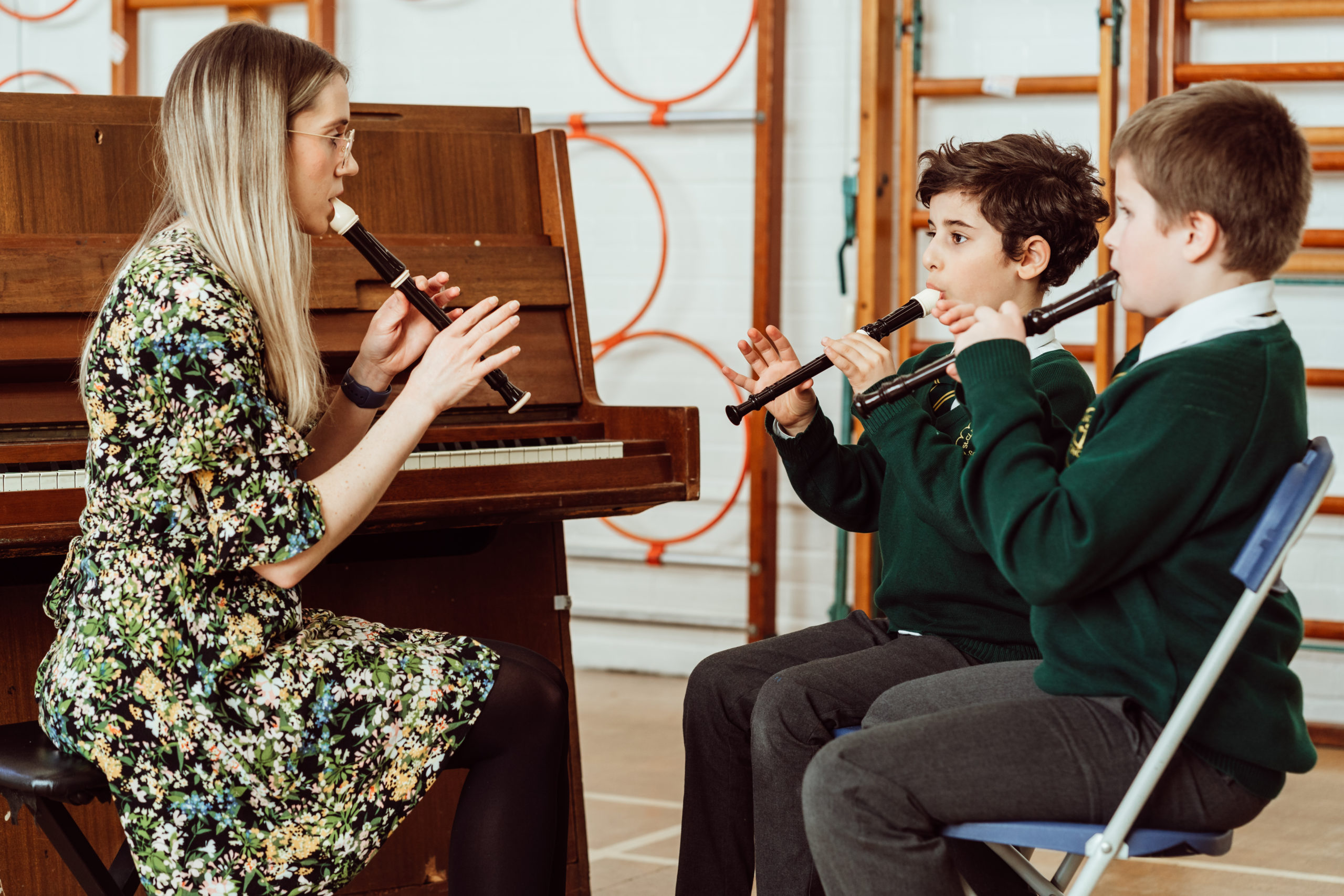 A music instructor in a floral dress sits by a piano, teaching two focused students in green school uniforms how to play the recorder. The trio, immersed in their lesson, exemplifies the hands-on, interactive approach to music education, fostering a harmonious learning environment.