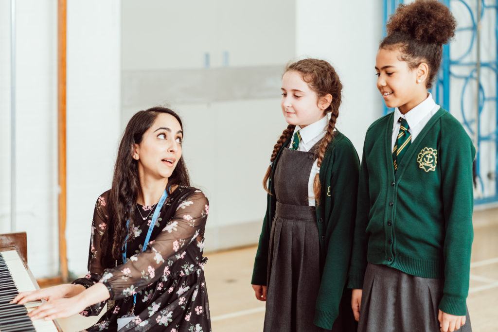 A music instructor, engaged and expressive, plays the piano and looks up at two attentive students in school uniforms, fostering a lively and interactive learning environment. The students, one with braided hair and the other with a bun, stand side by side, watching the teacher with interest, embodying the enriching experience of music education.