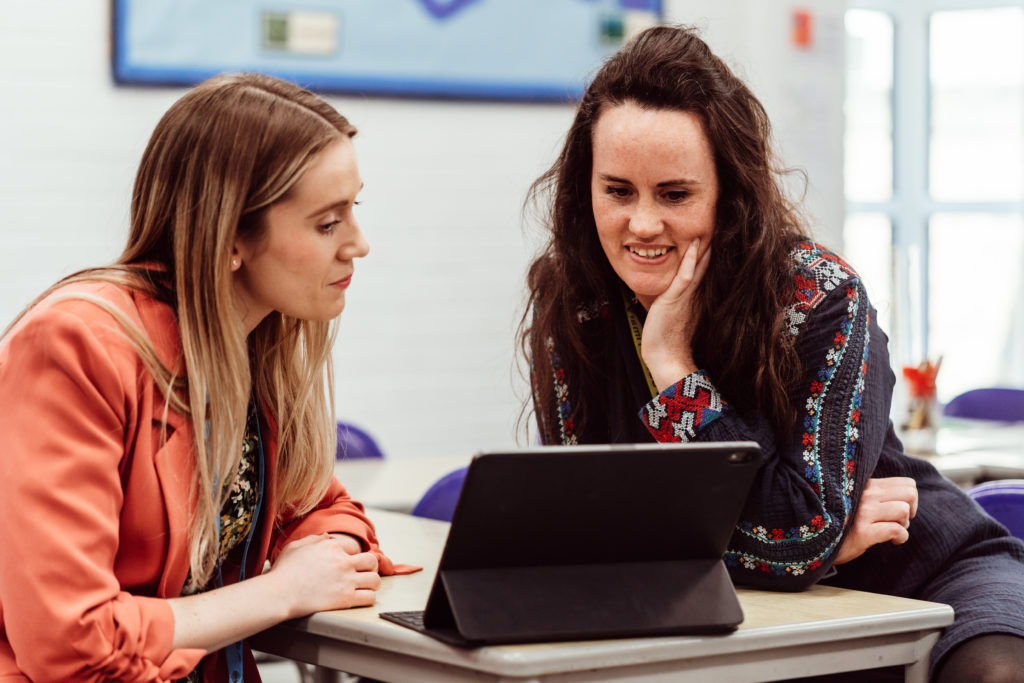 Two women are engaged in a collaborative activity, with one looking intently at a tablet screen while the other, wearing a lanyard, observes with interest. They are seated at a desk in a classroom environment, suggesting a focus on educational development or planning. The atmosphere is one of concentration and shared purpose, indicative of a productive educational setting.