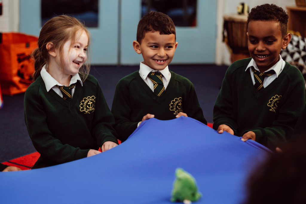Three young students in school uniforms share a moment of joy and laughter while engaging in a classroom activity with a blue fabric, with a small green plush toy on top, likely part of a learning game or exercise. Their bright smiles and the dynamic interaction capture the essence of a fun and collaborative educational experience.