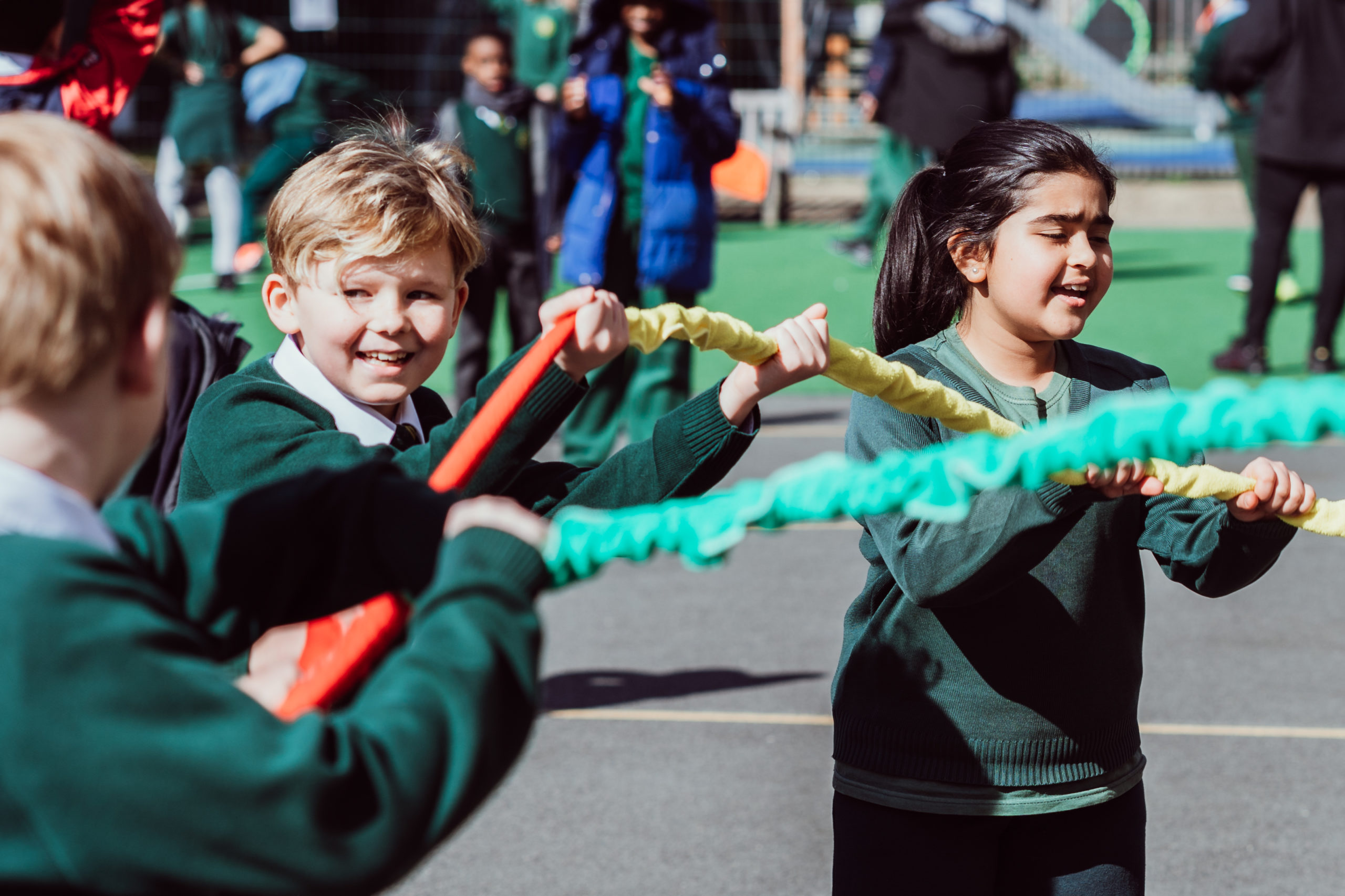 Children in school uniforms are engaged in a playful tug-of-war game under the bright sun, with expressions of joy and determination on their faces. The scene is filled with the energy of childhood and the spirit of friendly competition, set against the backdrop of a bustling schoolyard.