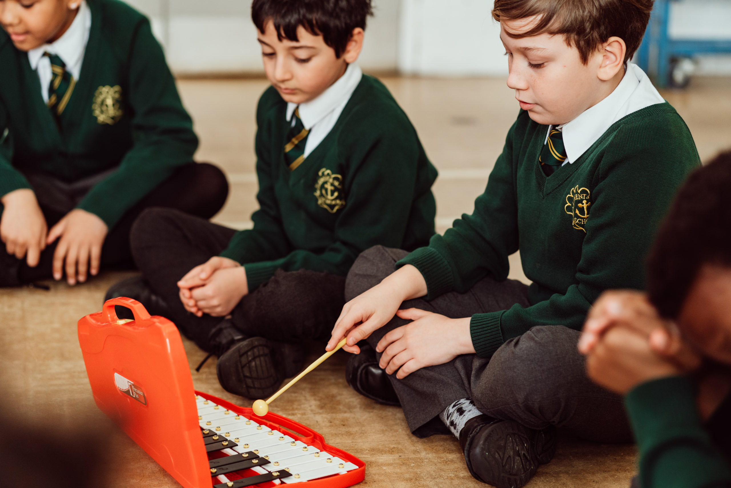 A focused child in a school uniform is seated on the floor, gently playing a colorful xylophone with a mallet, surrounded by attentive classmates who are observing and waiting for their turn, fostering a sense of musical exploration and peer learning in a classroom setting.