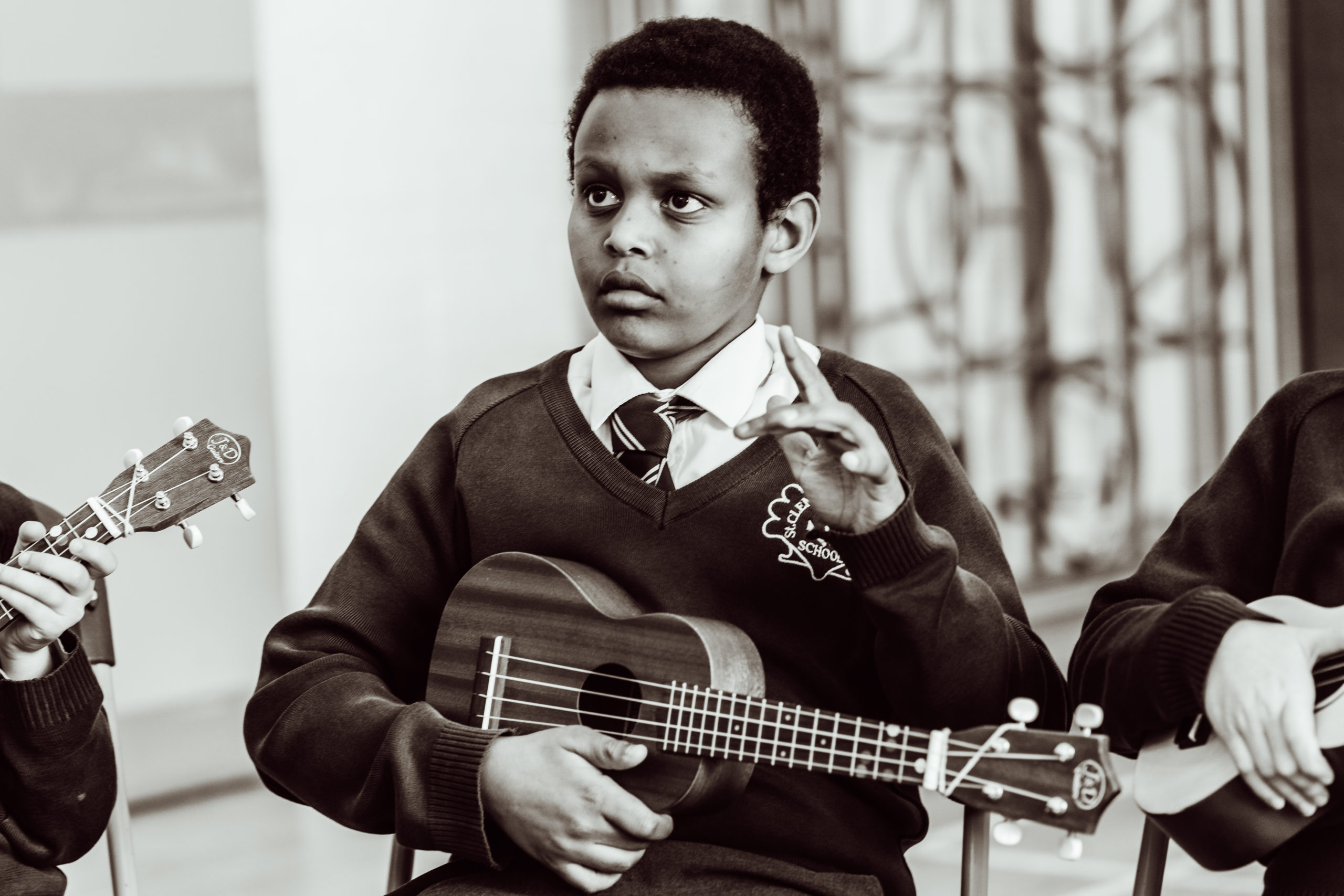 A focused schoolboy in a uniform holds a ukulele, attentively practicing his musical skills during a lesson, with a hint of concentration on his face as he learns to play the instrument.