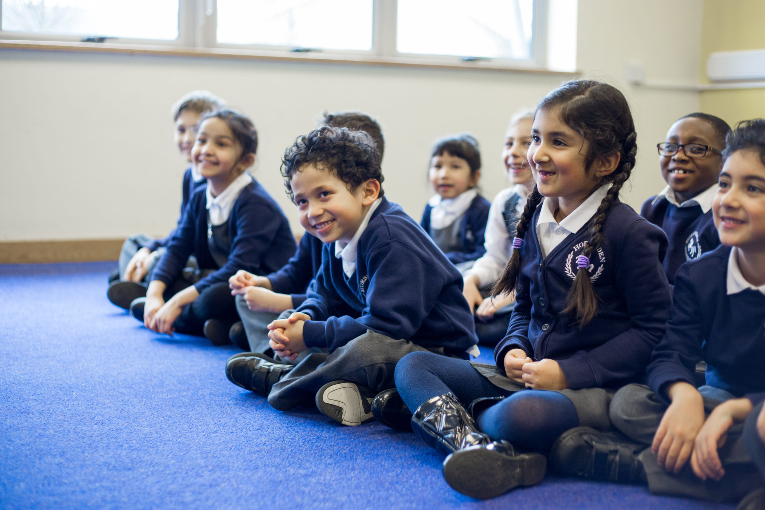 A group of cheerful young students in school uniforms sits on a blue carpet, their faces lit up with bright smiles as they engage in a classroom activity, exuding a sense of excitement and camaraderie.
