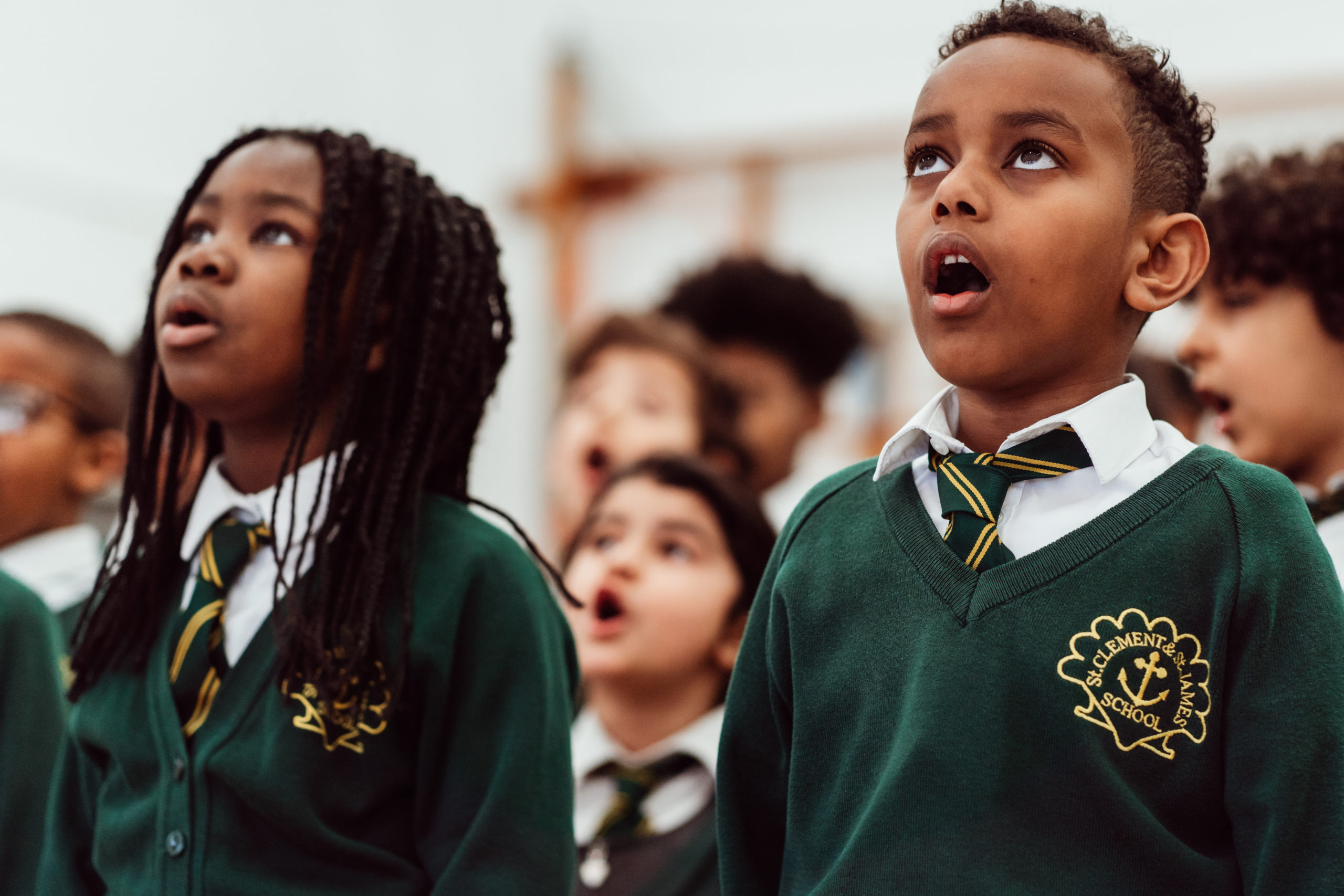 A group of schoolchildren, wearing green sweaters with school crests, are singing with expressions of focus and enjoyment. The children are engaged in a choral activity, showcasing the enriching experience of collective music-making in an educational setting.