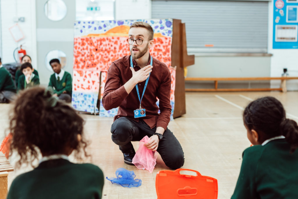 A captivated classroom watches an instructor, who is sitting cross-legged on the floor, demonstrating a lesson with colorful scarves, his expression animated and engaging, fostering a lively and interactive learning environment.
