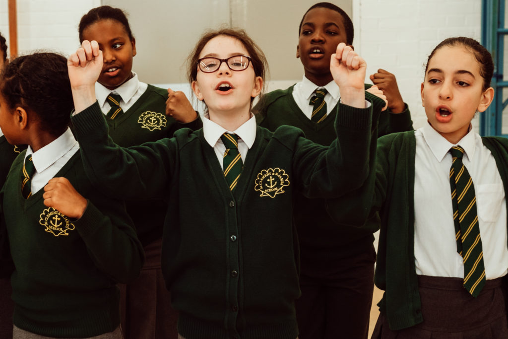 A group of enthusiastic schoolchildren in green uniforms with striped ties are actively participating in a group activity, singing or chanting with animated expressions and raised fists, demonstrating a sense of unity and engagement in their school environment.