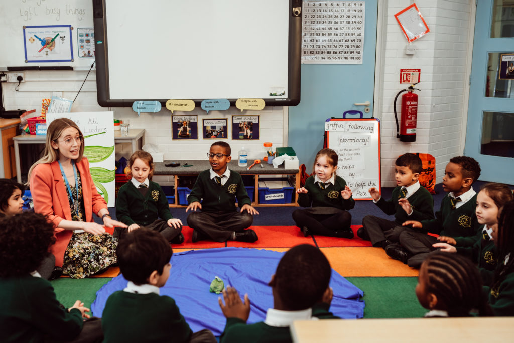 A vibrant classroom scene unfolds with a teacher engaging a circle of young students, who are attentively seated on colorful mats. The teacher, animated and expressive, leads an interactive lesson, while the children, dressed in green school uniforms, participate with enthusiasm. The classroom is adorned with educational materials and charts, creating an atmosphere of structured learning and active participation.