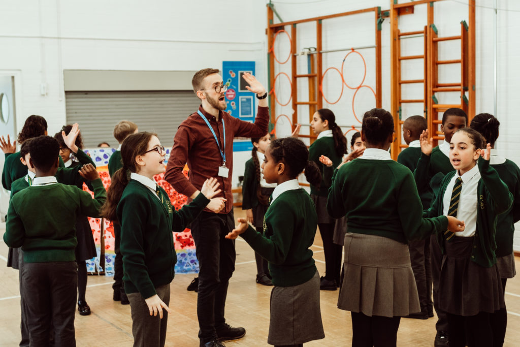 A lively music class is in session with a group of enthusiastic students participating in a singing activity led by an animated instructor. The children, wearing school uniforms, are spread out across the gymnasium, fully engaged in the exercise, with some raising their hands high and others gesturing as they sing. The atmosphere is one of joy and active learning, with the instructor at the heart of the action, guiding the students through the musical experience.