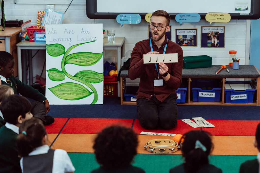 A music instructor sits cross-legged on a colorful classroom rug, engaging young students with a demonstration of musical notes using a handheld instrument. The children, seated attentively in a semi-circle, watch with interest. Behind the teacher, a poster reads "We Are All Learning Leaders," reinforcing a positive educational message amidst a setting equipped with learning aids and instruments, fostering a vibrant and interactive learning environment.