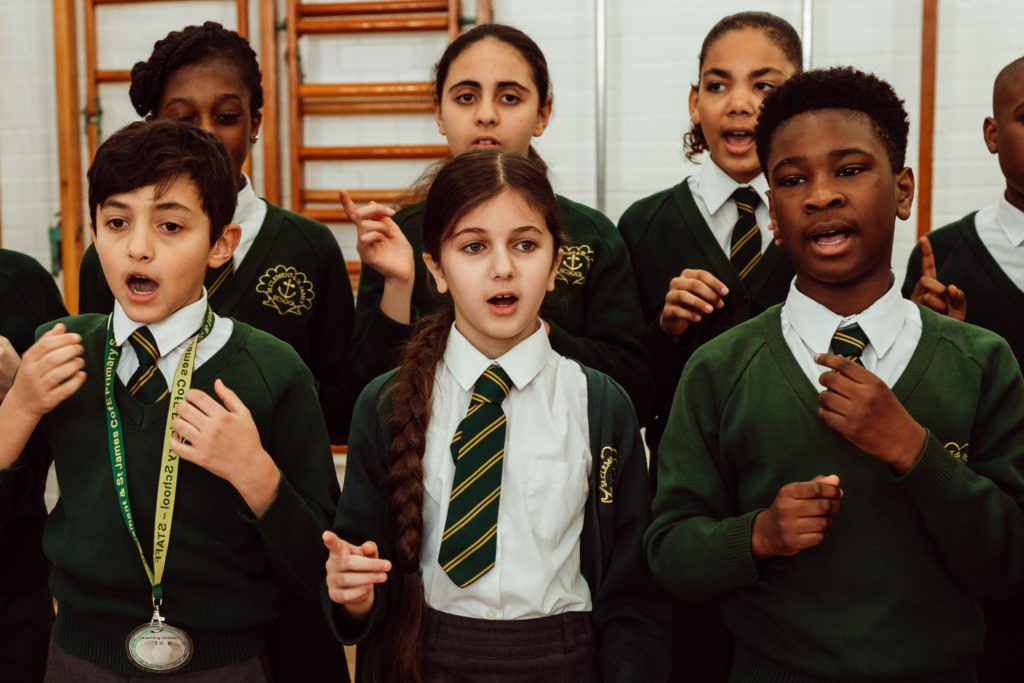 A group of enthusiastic schoolchildren, wearing green sweaters and striped ties as part of their uniform, are captured mid-song, with expressions of concentration and joy. Their hands gesture in rhythm, suggesting a lively and immersive music class experience that highlights the power of choral singing in education.