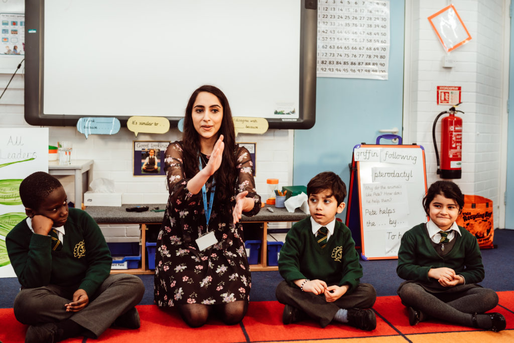 A teacher is engaging with young students seated on the floor in a colorful classroom. She gestures with her hands, illustrating a point, while the children, wearing school uniforms, look on with varying degrees of attention and curiosity. The background features educational materials and a whiteboard, creating an interactive learning environment.