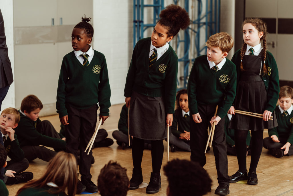 A group of schoolchildren in green sweaters and dark trousers or skirts are gathered in a school hall, some sitting and others standing attentively. Two standing children hold drumsticks, suggesting a music class or assembly, with a focus on participation and learning. The atmosphere is one of anticipation and concentration, as they seem to be waiting for instructions or the next part of their activity.