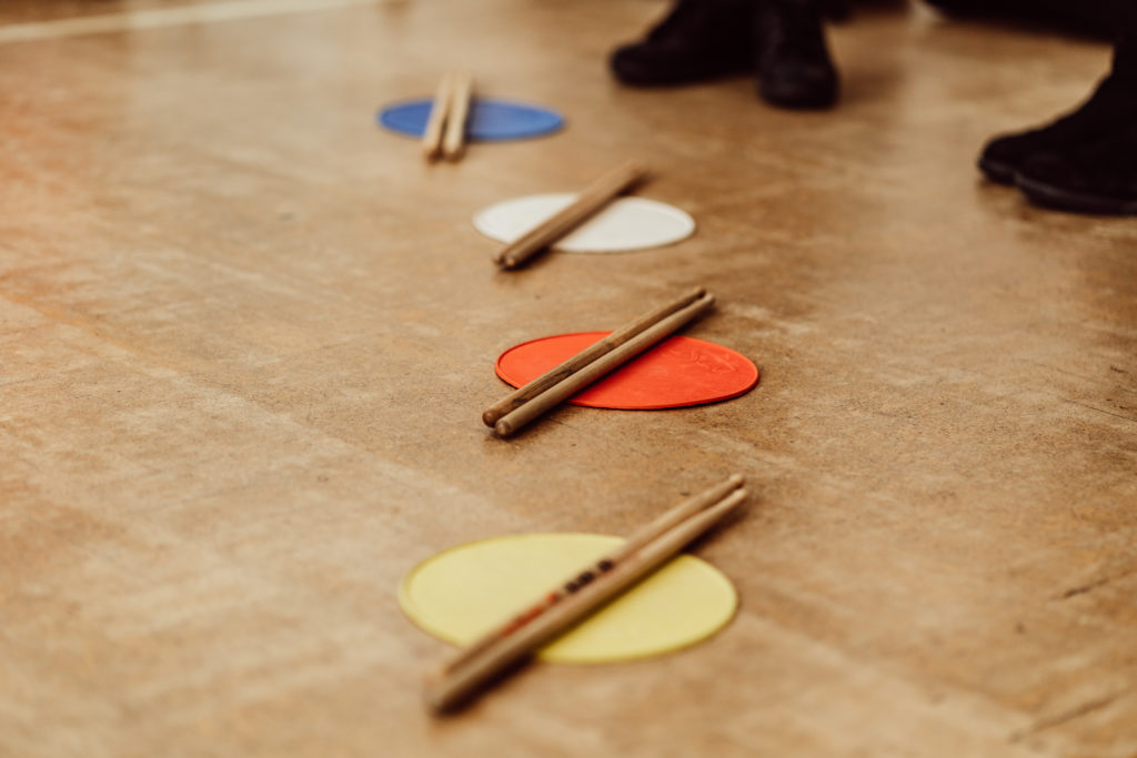 A close-up view of a wooden floor with colorful round markers in blue, white, red, and yellow, each with a pair of wooden drumsticks resting on top, suggesting a music class activity. In the background, the lower part of two individuals can be seen, indicating a social or educational setting.