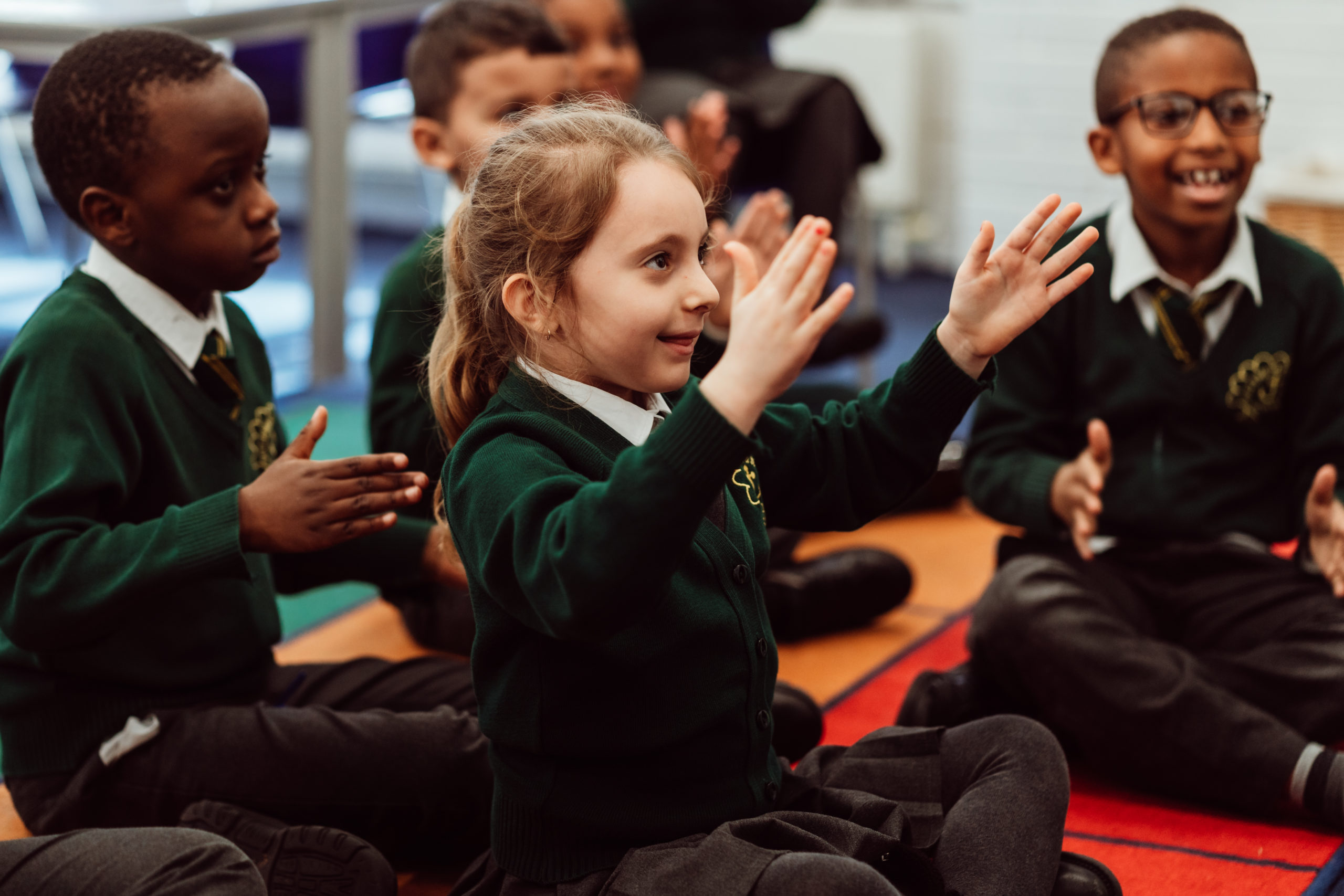 A group of schoolchildren in green uniforms are sitting on the floor, engaging enthusiastically in a clapping activity. The focus is on a young girl with a bright smile, clapping her hands with visible excitement, while her classmates around her participate with similar joy and concentration. The atmosphere is lively and suggests a fun, interactive learning environment.