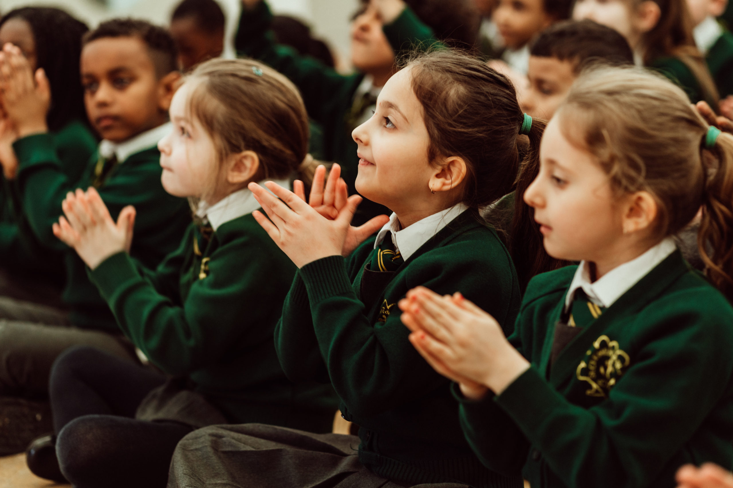 A group of young students, wearing green school uniforms, are seated with their attention focused to the left, possibly watching a performance or listening to a teacher. Their hands are clapped together in a gesture that suggests they are engaged in a learning activity or applauding, with expressions of interest and enjoyment on their faces. The atmosphere is one of attentive participation and collective experience in an educational setting.