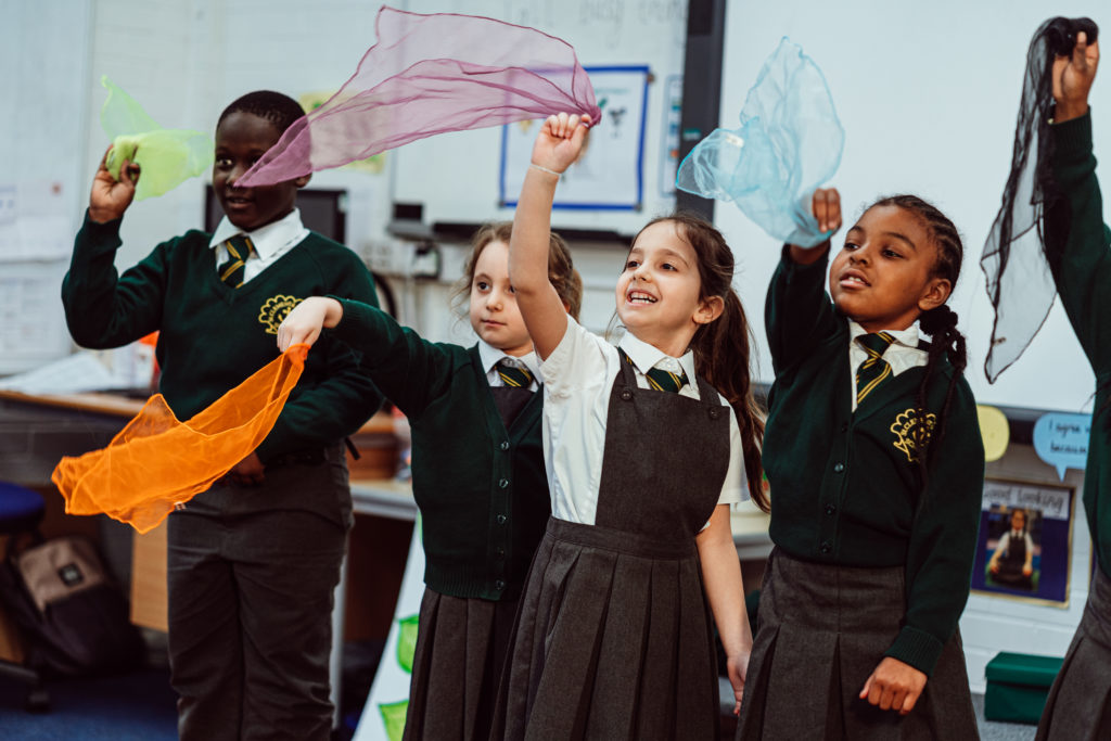 A group of joyful schoolchildren, wearing green sweaters with school crests, are engaged in a classroom activity, waving colorful sheer scarves in the air. Their expressions are bright and animated, capturing a moment of playful learning and creative expression.