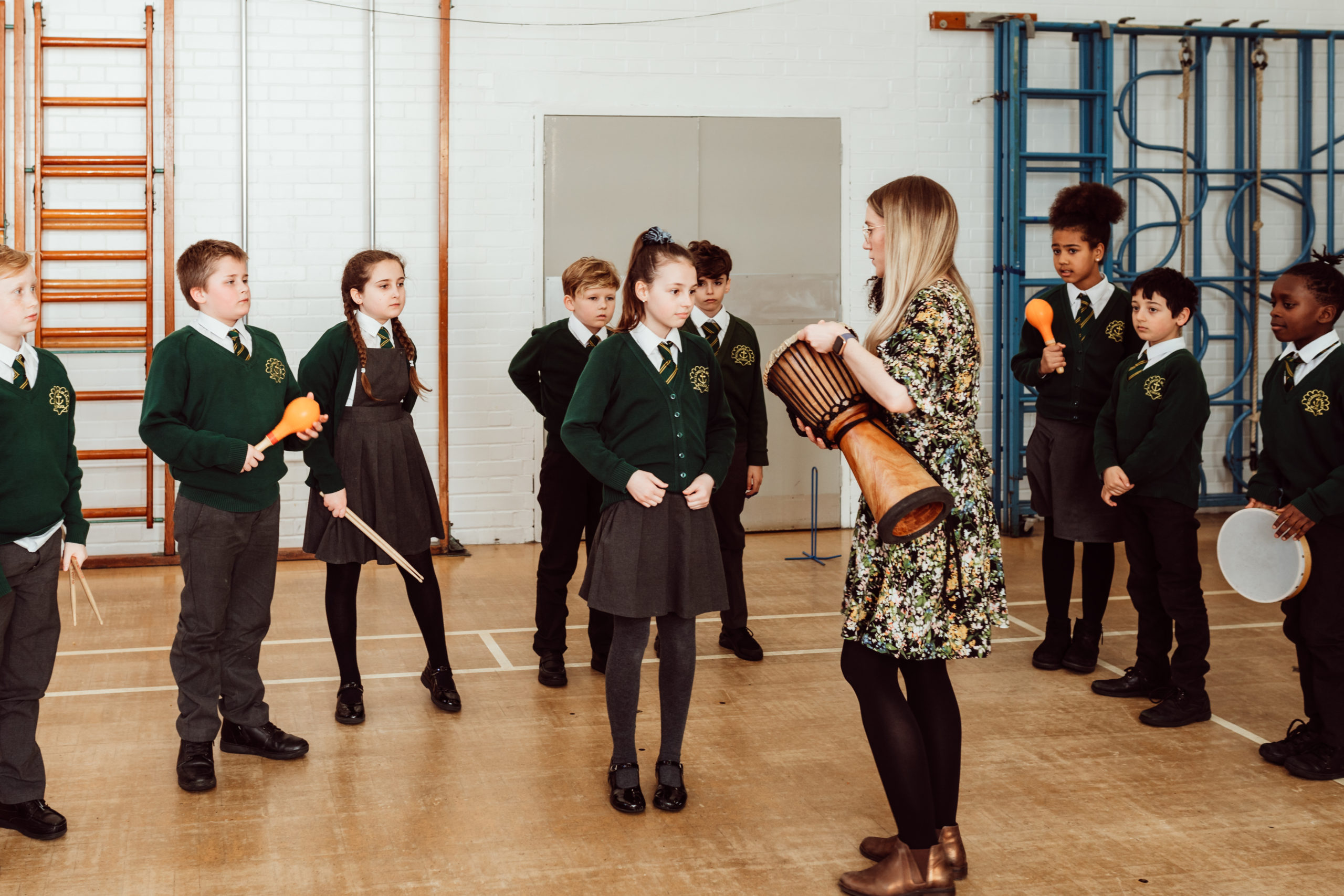 A group of attentive schoolchildren, clad in green sweaters and dark bottoms, stand in a gymnasium, focusing on a teacher who is demonstrating how to play a wooden djembe drum. Some students hold percussion instruments like maracas and tambourines, ready to participate in the music lesson, creating an atmosphere of interactive learning and cultural exploration.