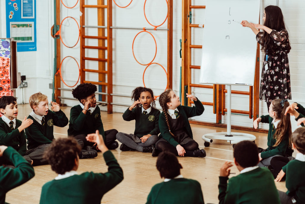 A group of schoolchildren in green uniforms are seated on the floor, attentively participating in a music lesson led by a teacher who is writing on a whiteboard. The children appear engaged and animated, some with hands raised, mimicking musical notes or gestures, in a bright classroom with climbing bars on the wall, indicating a lively and interactive learning environment.