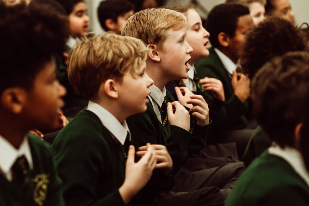 A group of schoolchildren, dressed in green uniforms, are captured in a moment of collective focus, their expressions reflecting engagement and enthusiasm. They appear to be singing or participating in a vocal activity, with some children gesturing as if emphasizing the rhythm or melody of a song they are learning together. The atmosphere suggests a lively and harmonious learning experience.