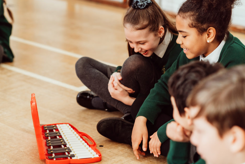A group of schoolchildren, wearing green sweaters and sitting on the floor, are gathered around a red glockenspiel, sharing a moment of laughter and learning. The focus is on two girls in the foreground, one with a joyful smile, as they engage in a music education activity, highlighting the camaraderie and interactive nature of the lesson.