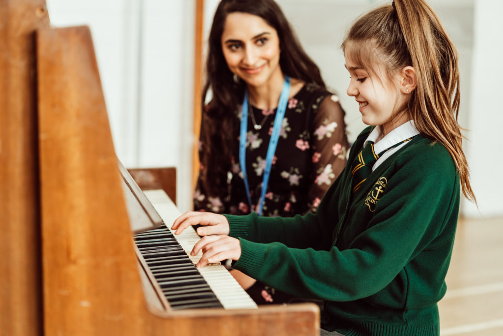 A young student in a green school sweater is playing the piano, focused on the keys, while a smiling teacher with a lanyard stands beside her, observing and offering guidance, creating an atmosphere of learning and encouragement in music education.