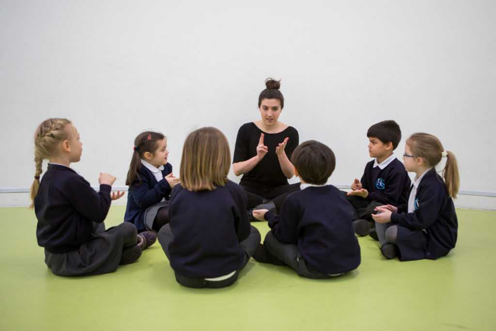 A teacher sits on the floor with a circle of young students, all engaged in a lively music lesson. The children, wearing school uniforms, appear focused and animated, some with hands mid-gesture, suggesting they are either singing or participating in a rhythmic activity. The teacher, central to the group, guides the activity with expressive hand movements, fostering an atmosphere of learning through interactive participation.