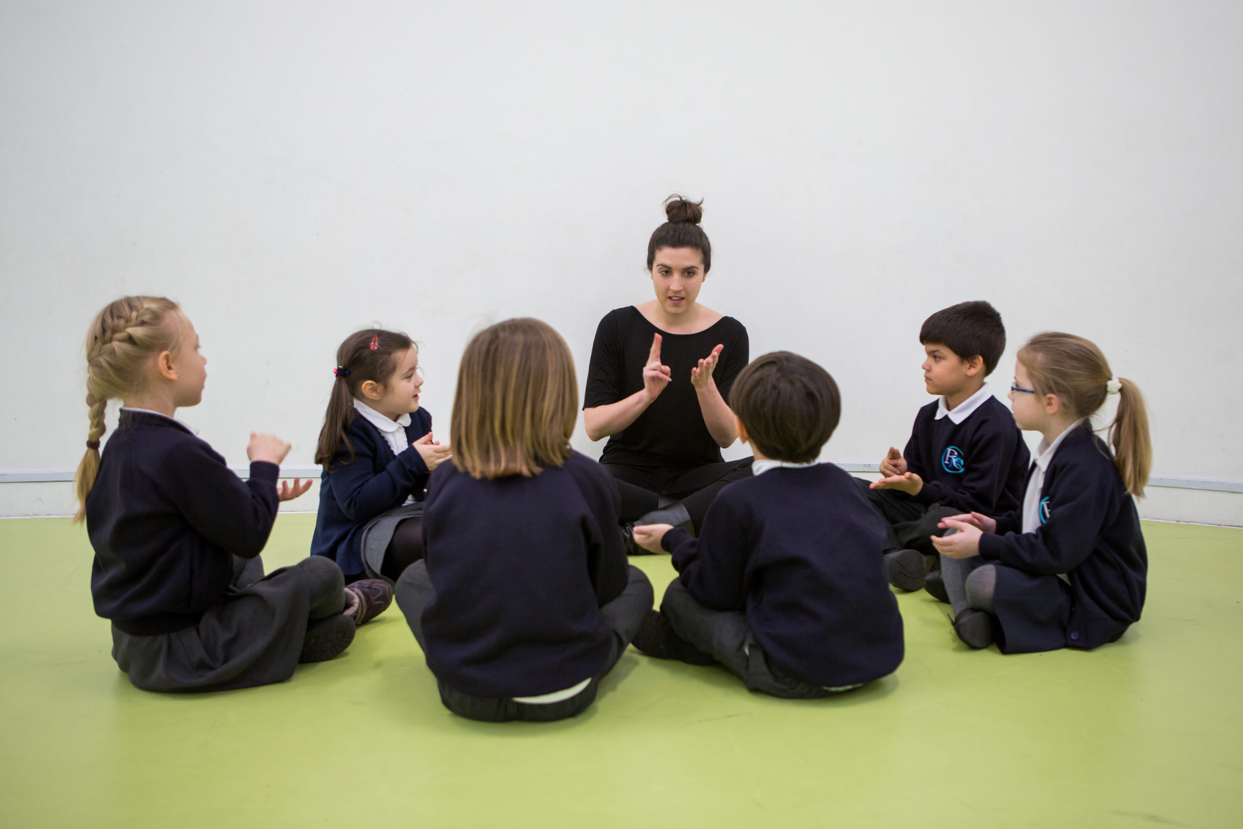 A teacher sits on the floor with a circle of young students, all engaged in a lively music lesson. The children, wearing school uniforms, appear focused and animated, some with hands mid-gesture, suggesting they are either singing or participating in a rhythmic activity. The teacher, central to the group, guides the activity with expressive hand movements, fostering an atmosphere of learning through interactive participation.